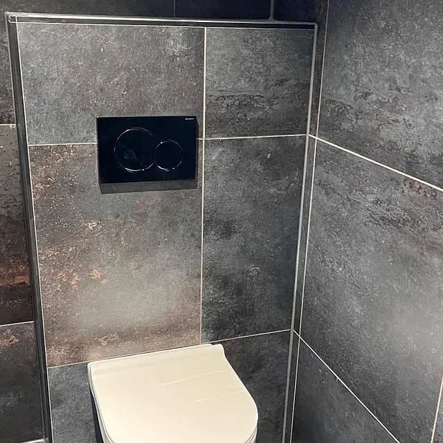 Vegghengt toalett med sort trykkplate montert på vegg med mørke, store fliser som gir et stilrent og moderne uttrykk.