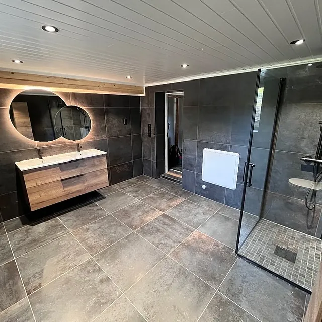 Romslig bad med store mørke fliser, dusjhjørne i glass, dobbel servant med opplyste speil og moderne innredning.