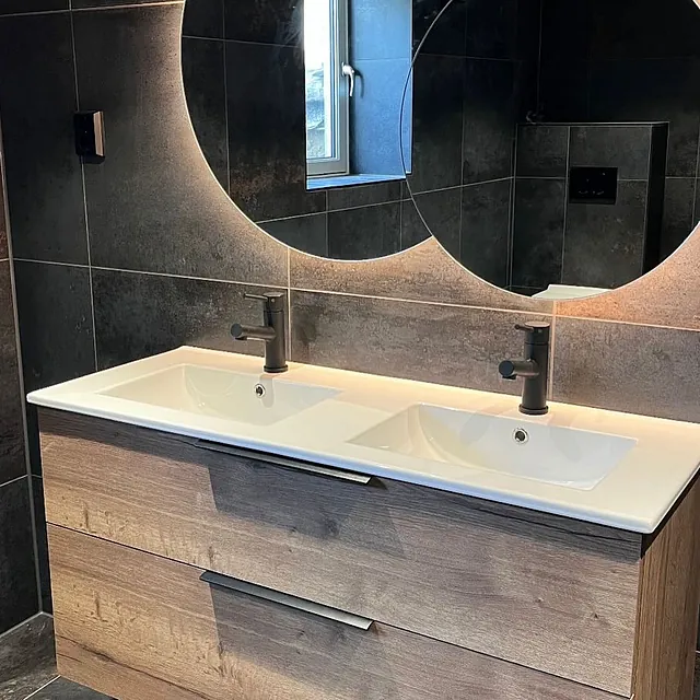 Dobbel servant på baderomsmøbel i tre med sorte armaturer og runde speil med integrert belysning mot mørke fliser.