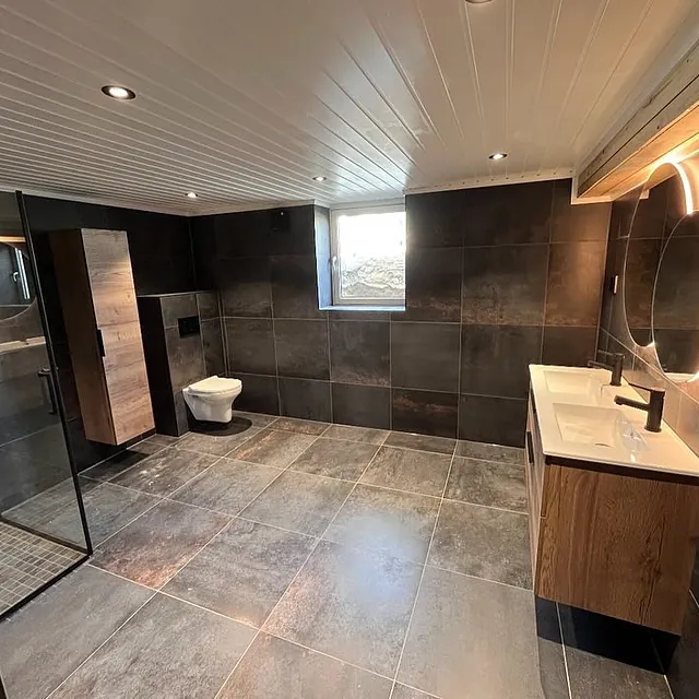 Stort moderne bad med dusjhjørne i glass, vegghengt toalett, mørke fliser og servant med rundt speil og stemningslys.